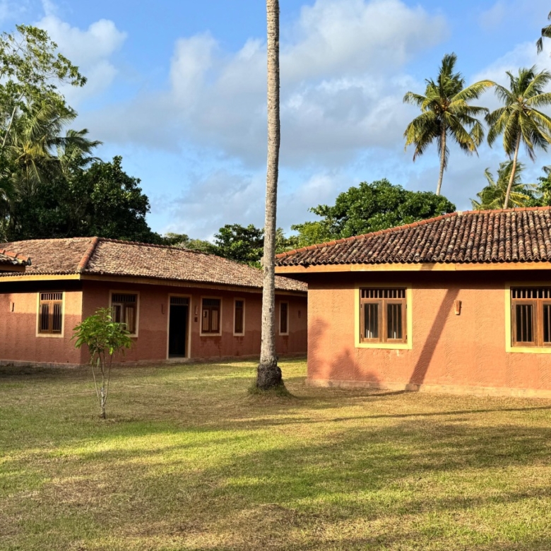 Terracotta-colored buildings with red-tiled roofs in a tropical courtyard among palm trees under a blue sky.