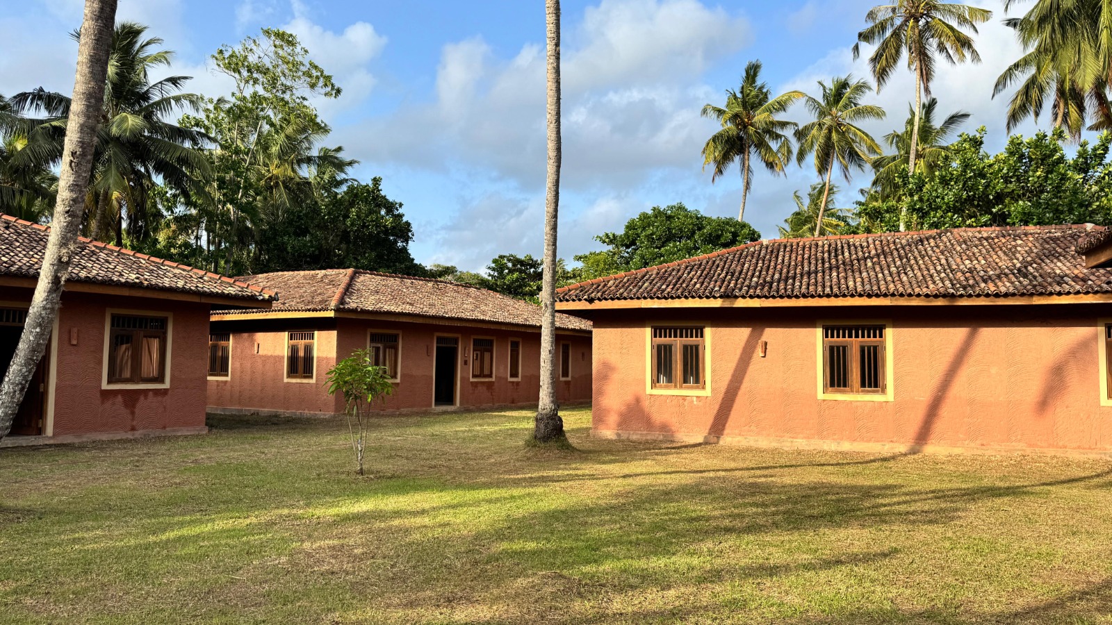 Terracotta-colored buildings with red-tiled roofs in a tropical courtyard among palm trees under a blue sky.